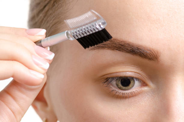 A close-up image of a person grooming their eyebrow with a small brush. The focus is on the details of the eyebrow and the tool being used for styling.