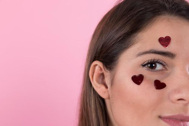 Close-up of a woman with heart stickers on her face against a pink background